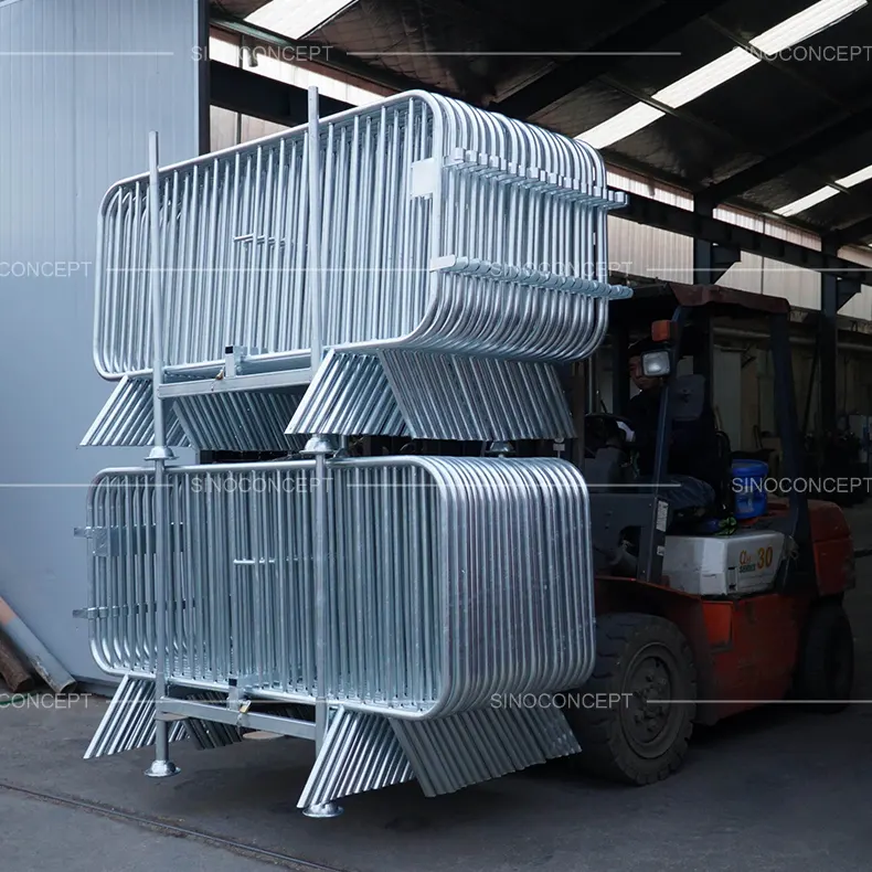 Forklift lifting double-stacked galvanised steel crowd barrier stillages loaded with metal barriers, showing stable stackable design for bulk storage and transport