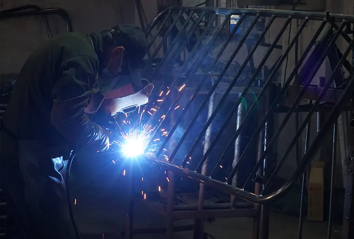 A worker welding a crowd control barrier, with visible sparks, in a manufacturing facility